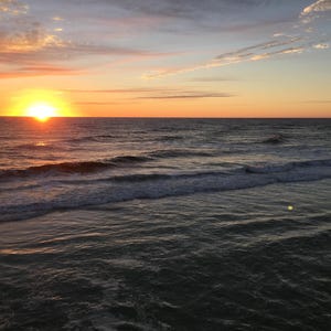 May include: A sunset over the ocean with waves breaking on the shore. The sky is a mix of orange, pink, and purple.