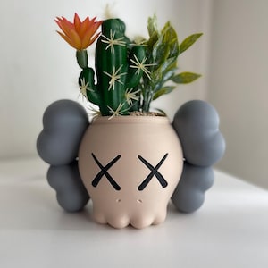 May include: A light pink ceramic planter shaped like a cartoon character with gray ears and black X eyes. The planter is filled with a green cactus and a small green plant with a single orange flower.