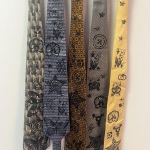 Puede incluir: Cinco corbatas colgadas una al lado de la otra, cada una con un patrón base único y diseños estampados en tinta negra que incluyen mariposas, estrellas, flores y formas geométricas. Las corbatas son de tonos beige, gris, dorado y amarillo.