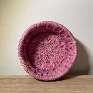 May include: A pink woven basket made of straw. The basket is round and has a woven pattern.