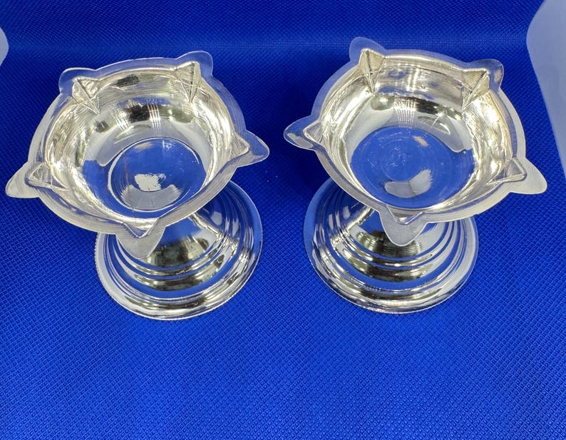 Op de afbeelding: Twee zilveren diya-olielampen met een stervormig ontwerp. De lampen staan op een blauw oppervlak.