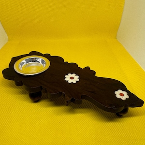 May include: A dark brown wooden decorative tray with a silver-colored bowl and three white flower accents with red centers. The tray has a unique, curved shape and rests on small wooden feet.