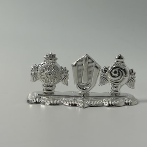 May include: A silver-colored religious artifact with three ornate components. The central piece is a shield-like shape, flanked by two decorative, circular elements. The base is a scalloped design. The object is set against a plain white background.