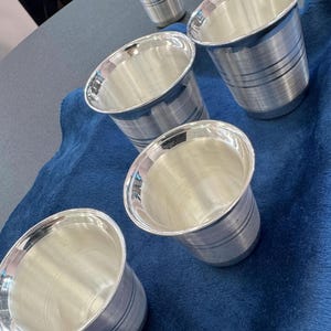 May include: A set of four silver-colored metal tumblers with a polished finish and horizontal line detailing around the exterior. Each tumbler features a wide, flared rim and a tapered base, arranged in a cluster to show their reflective surfaces.