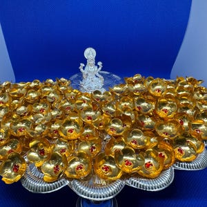 May include: A silver-toned pedestal tray holds a large collection of small, gold-coloured flower-shaped ornaments, each featuring a single red rhinestone at its centre. A small, silver-toned figurine of a deity stands at the back of the tray.
