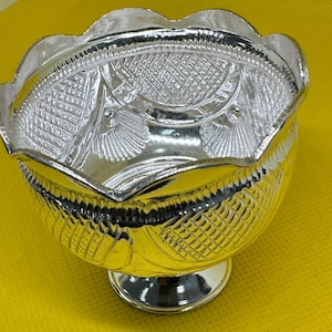 May include: A silver bowl with a decorative, scalloped edge and a pedestal base. The bowl's interior has detailed geometric patterns and shell motifs. The bowl is made of a shiny, reflective material.