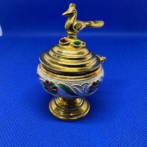 Op de afbeelding: Een gouden en witte metalen wierookbrander met een pauwenkop. De brander heeft een ronde basis en een deksel met een pauwenontwerp. De pauw is groen, goud en rood.
