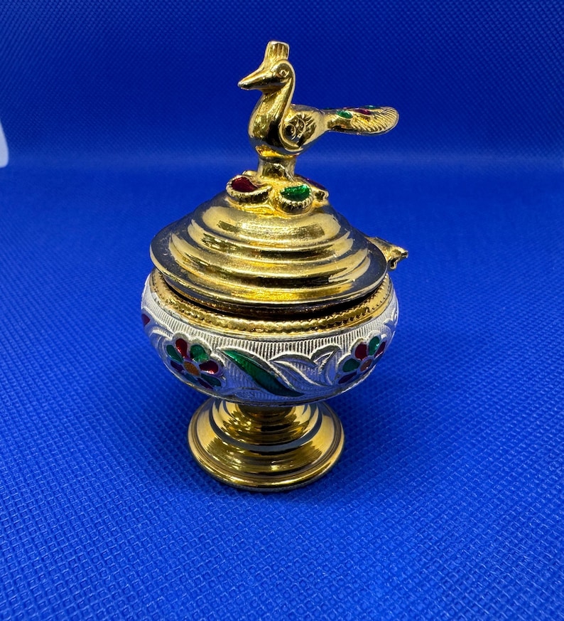 Op de afbeelding: Een gouden en witte metalen wierookbrander met een pauwenkop. De brander heeft een ronde basis en een deksel met een pauwenontwerp. De pauw is groen, goud en rood.