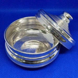 Op de afbeelding: Een ronde, zilveren container met een deksel en een glad, reflecterend oppervlak. Het deksel heeft een kleine knop aan de bovenkant.