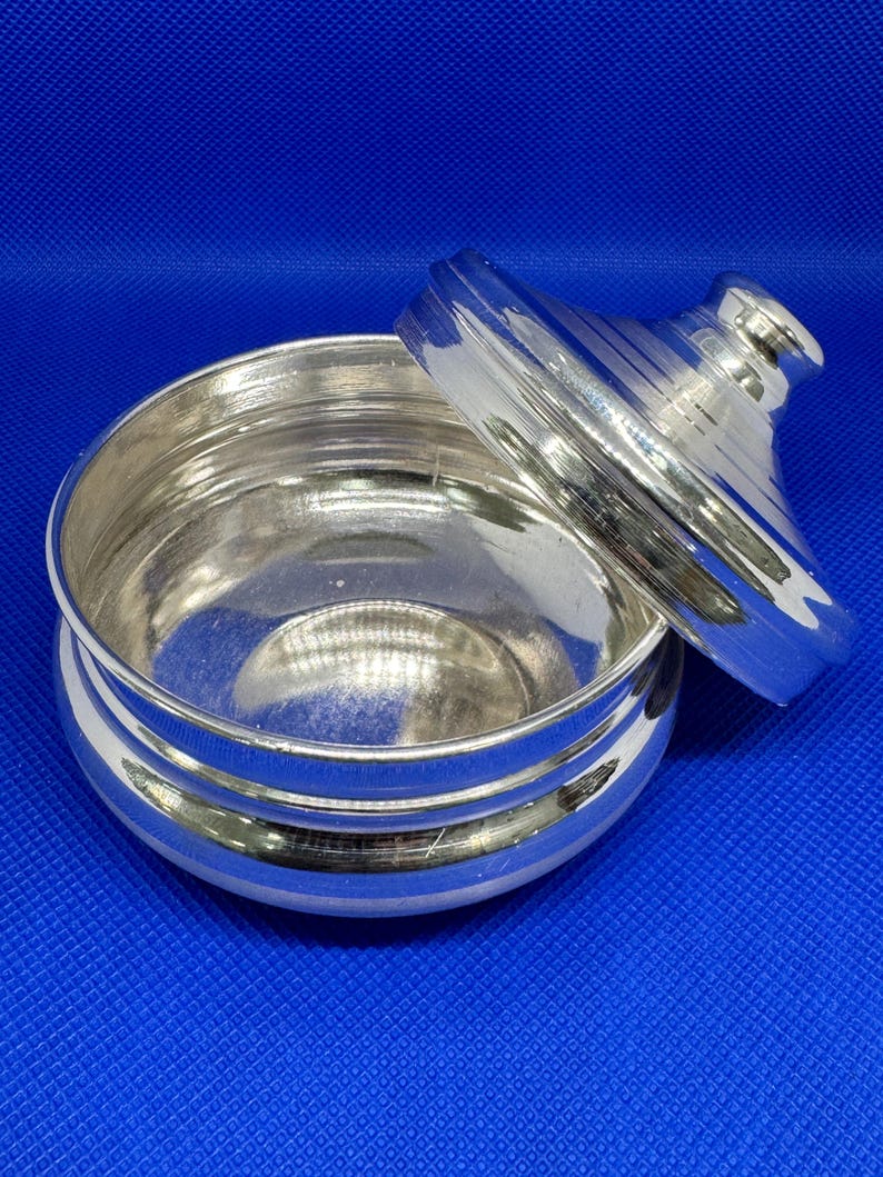 Op de afbeelding: Een ronde, zilveren container met een deksel en een glad, reflecterend oppervlak. Het deksel heeft een kleine knop aan de bovenkant.