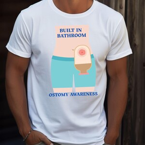 May include: White t-shirt with a graphic design promoting ostomy awareness. The design features an illustration of a person with a stoma and the words "BUILT IN BATHROOM" and "OSTOMY AWARENESS" in blue text.