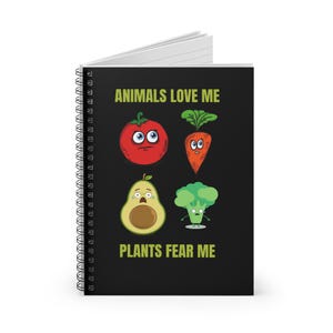 May include: A spiral-bound notebook with a black cover. The cover displays the text "ANIMALS LOVE ME" and "PLANTS FEAR ME", alongside cartoon illustrations of a tomato, carrot, avocado, and broccoli, each with expressive faces.
