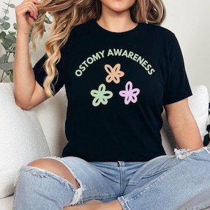 May include: Black t-shirt with the text "Ostomy Awareness" in a white arc above three colorful flower illustrations.
