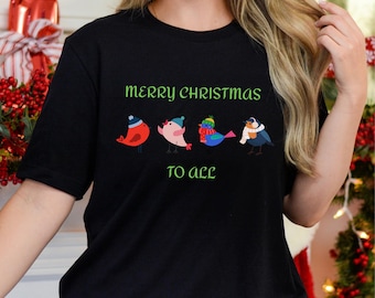 Könnte beinhalten: Schwarzes T-Shirt mit dem grünen Text "MERRY CHRISTMAS TO ALL". Das Shirt zeigt vier Cartoon-Vögel mit Winteraccessoires. Die Vögel sind rot, rosa, blau und grau, mit Mützen und Schals.