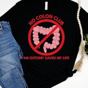 May include: Black t-shirt with the text "NO COLON CLUB" in red, with a pink colon image crossed out. The text "AN OSTOMY SAVED MY LIFE" is below the image. The shirt is paired with blue jeans.