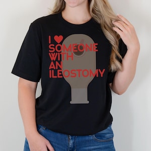 May include: Black t-shirt with the text "I ❤️ SOMEONE WITH AN ILEOSTOMY" in red. The design includes a red heart and a graphic illustration of an ileostomy bag. The shirt is a casual, short-sleeved style.