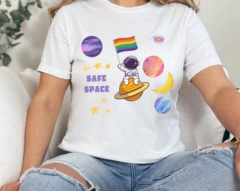 May include: White t-shirt featuring a graphic of an astronaut holding a rainbow flag in space. The text "SAFE SPACE" is printed on the shirt.