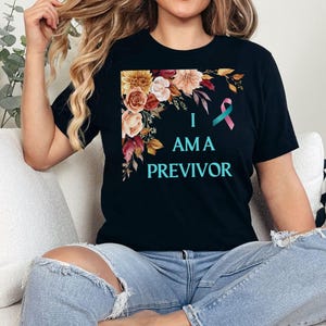 May include: Black t-shirt with a floral design in the corner and a teal and pink ribbon. The text "I am a Previvor" is printed on the shirt.