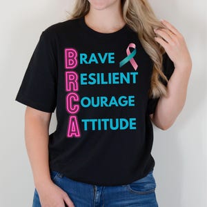 May include: Black t-shirt with the text "BRAVE RESILIENT COURAGE ATTITUDE" in teal and pink neon lettering. A teal and pink ribbon is also featured on the shirt. The shirt is a crew neck style.