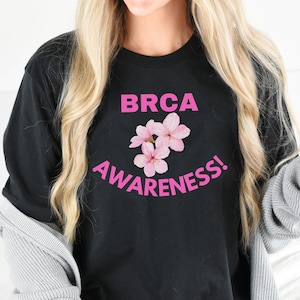 May include: Black t-shirt with pink text that reads "BRCA AWARENESS!" and pink flower graphic.