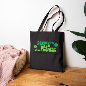 May include: Black tote bag with the words "PREVIVOR BRCA AWARENESS" in green, with flower accents. The bag has black handles and is likely made of canvas or a similar material. The bag is sitting on a wooden surface.