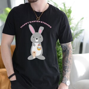 May include: Black t-shirt with a gray bunny holding a basket of Easter eggs. The text "Happy Easter Stomies" is printed in pink above the bunny.