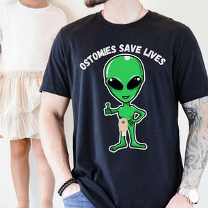 May include: Black t-shirt with the text "OSTOMIES SAVE LIVES" above a green alien illustration. The alien has a thumbs-up gesture and a beige ostomy bag. A child in a white top and skirt is in the background.
