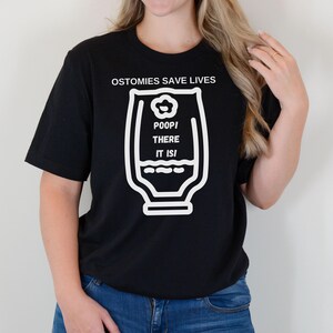 May include: Black t-shirt with white text and graphic. The text reads "OSTOMIES SAVE LIVES" above a graphic of a medical bag with the words "POOP! THERE IT IS!" inside. The shirt is paired with blue jeans.