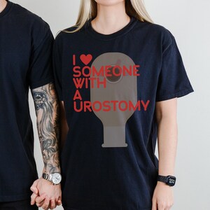 May include: Black t-shirt with the text "I ❤️ SOMEONE WITH A UROSTOMY" in red. The design includes a red heart and a graphic of a urostomy bag. The shirt is worn by a person.