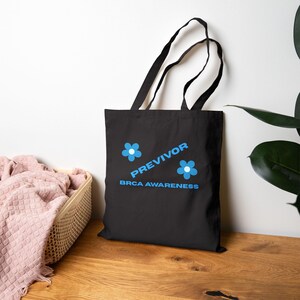May include: Black tote bag with blue text and flower graphics. The text reads "PREVIVOR BRCA AWARENESS". The bag has two black handles and is made of a sturdy material. The design is centered on the front of the bag.
