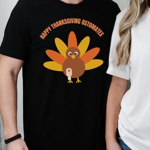 May include: Black t-shirt with a cartoon turkey graphic. The turkey has brown, orange, and yellow feathers, and is holding a beer. The text "HAPPY THANKSGIVING OSTOMATES" is arched above the turkey.