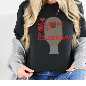 May include: Black t-shirt with the text "I love someone with a colostomy" in red, along with a red heart graphic. The shirt features a graphic of a colostomy bag.