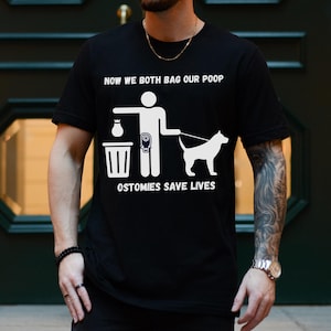 May include: A black t-shirt with a white graphic of a person and a dog both holding bags. The text on the shirt reads "Now we both bag our poop" and "Ostomies save lives".
