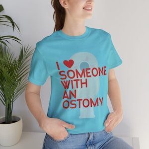 May include: A light blue t-shirt with the text "I ❤️ SOMEONE WITH AN OSTOMY" in red. The design includes a red heart and a stylized illustration of an ostomy appliance. The shirt is made of a soft material.