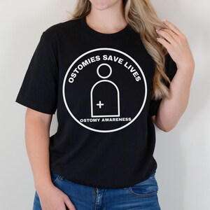 May include: A black t-shirt with a white circle graphic that says "Ostomies Save Lives" and has a stylized person with a plus sign inside. The text "Ostomy Awareness" is below the circle.