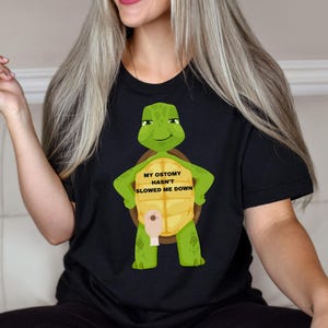 May include: Black t-shirt featuring a cartoon turtle with the text "MY OSTOMY HASN'T SLOWED ME DOWN". The turtle is green and yellow, with a smiling face and arms. The shirt is made of a soft material.