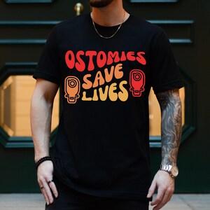 May include: Black t-shirt with the text "OSTOMIES SAVE LIVES" in red, orange, and yellow. The design includes two stylized illustrations. The shirt is worn by a person with a beard and tattoos.