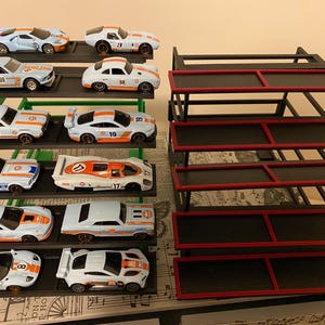 May include: A collection of twelve die-cast toy cars with Gulf livery, featuring blue and orange paint schemes, displayed on a green tiered stand. Beside it sits an empty black tiered display stand with red trim on each of the five shelves.