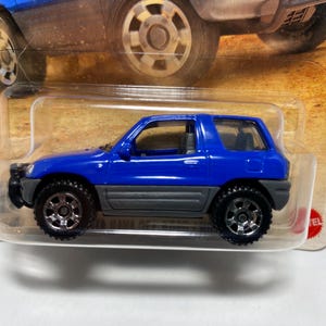 May include: A blue toy SUV with black wheels and a gray lower body. The toy car is in a clear plastic package, with a partial view of another car above it. The wheels have silver rims.