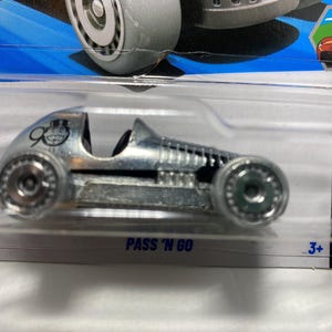 May include: A silver die-cast toy car with black accents, packaged in a clear plastic case. The car has large wheels and the text "PASS 'N GO" is visible. The packaging also includes the number "3+".