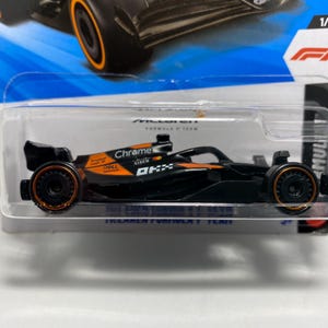 May include: A black and orange McLaren Formula 1 race car toy in a clear plastic package. The car has orange wheels and accents, with the words "Chrome" and "OKX" visible on the side. The packaging includes the F1 logo and the number "1/5".