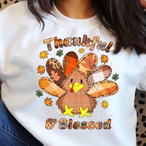 Könnte beinhalten: Weißes Sweatshirt mit einem Thanksgiving-Design. Die Grafik zeigt einen Truthahn mit karierten Federn und den Worten "Thankful & Blessed" in einer herbstlichen Schriftart, umgeben von Herbstblättern.