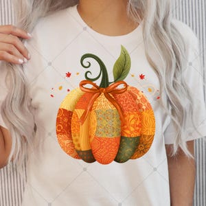 Puede incluir: Camiseta blanca con un diseño de calabaza de retazos coloridos. La calabaza tiene secciones naranjas, amarillas y verdes, un lazo decorativo y un tallo. Camiseta gráfica con temática otoñal.