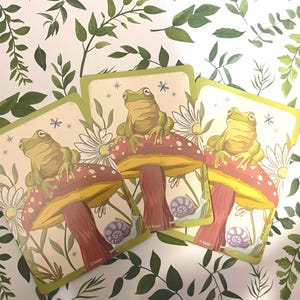 May include: Three cards featuring a whimsical illustration of a frog perched atop a red and yellow mushroom. The cards have a light green border and include a snail, daisies, and the text "Kalan". The background is a white surface with green leaves.