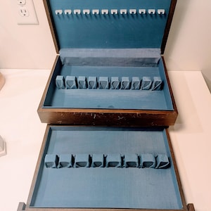 May include: A wooden silverware chest with a dark brown finish, open to reveal a blue velvet interior. The chest has two levels, each with slots for utensils. The top level has white plastic holders for knives. The chest is on a white surface.