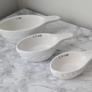May include: Three white ceramic measuring cups with a textured floral design. The cups are labeled "1 cup", "1/2 cup", and "1/4 cup" in black text. Each cup has a handle.