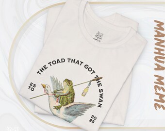 Könnte beinhalten: Cremefarbenes T-Shirt mit einer humorvollen Illustration einer grünen Kröte, die auf einem weißen Schwan reitet und eine Flasche an einer Angelrute hält. Der Text "THE TOAD THAT GOT THE SWAN" ist über dem Bild. Das Shirt hat auch den Text "ESTD 2025".