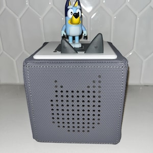 May include: A gray, cube-shaped speaker with a cat-shaped speaker grill. A small Bluey figurine stands on top, next to two gray shark fin-shaped buttons. The speaker is on a white surface against a white tile background.