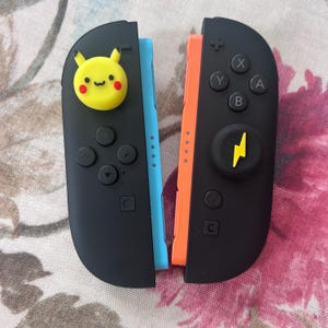 May include: Two black Nintendo Switch Joy-Con controllers, one with a yellow Pikachu face and the other with a yellow lightning bolt. The left controller has a blue side, and the right has an orange side. The controllers have buttons and directional pads.