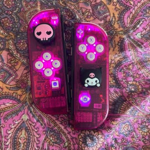 May include: Two pink Nintendo Switch Joy-Con controllers. One has a skull charm, the other a Kuromi charm. Both have illuminated buttons and directional pads. The controllers are set against a patterned pink and orange background.